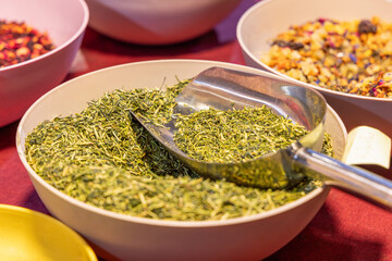 Bowl filled with loose green tea leaves with a metal scoop resting inside surrounded by other bowls containing various fruit tea blends in the background