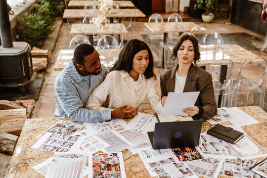 A woman and a man are sitting at a table and looking at a sheet of paper held by a woman sitting next to them, in an establishment, during wedding preparations