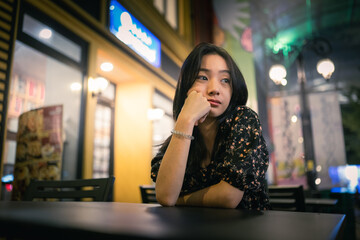 An Asian woman sits at a café table, resting her chin on her hand, appearing thoughtful. The warm indoor lights and colorful building facade create a cozy evening vibe