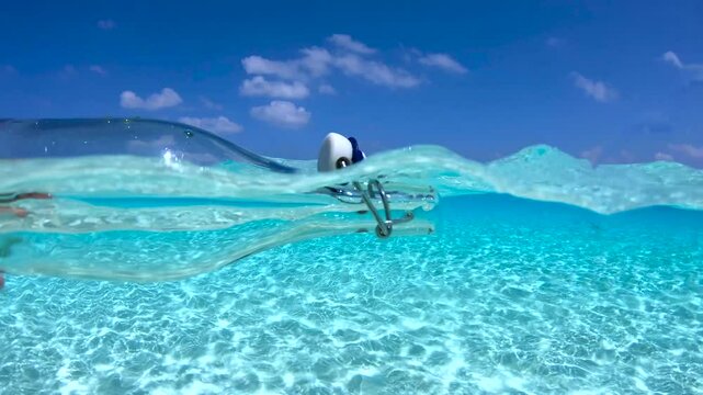 A hand reaches for a glass bottle submerged in the clear water of the Maldives, with a bright sky and scattered clouds visible above the surface.