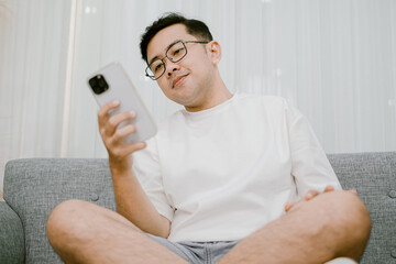 Southeast Asian man wearing glasses and a white t-shirt sitting on a couch using a smartphone