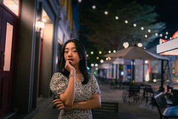An Asian woman in a floral dress stands pensively on a city street at night, with warm lights and a blurred background of shops and glowing bokeh