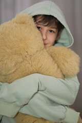 A boy in a pastel hoodie hugs a large fluffy teddy bear, looking shyly at the camera.