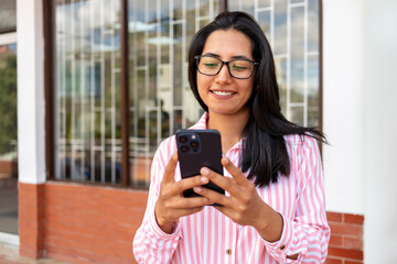 Smiling young Hispanic woman using her smartphone while walking outside office building