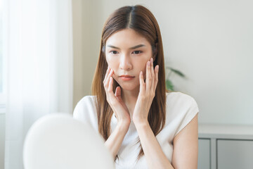 Anti aging, worry puberty asian young woman, stressed girl looking in mirror, allergy presenting an allergic reaction from cosmetic on face. Beauty, health care from acne problem, dry skin, treatment.