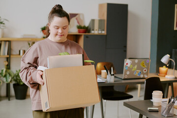 Wide shot of young woman with down syndrome carrying cardboard box with houseplant and notebooks in...