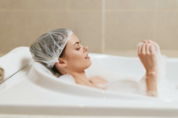 Relaxed young woman enjoying a bubble bath in a modern bathroom, wearing a shower cap and surrounded by soft white lather. Self-care and wellness concept.