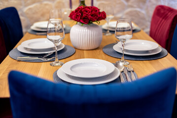 Elegant dining table set with white porcelain plates, wine glasses, and silverware, featuring a red rose centerpiece in a stylish white vase