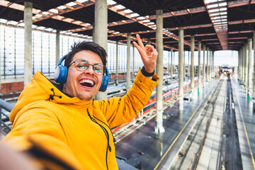 Happy traveler taking a selfie at a train station. Smiling man with headphones and a yellow jacket taking a selfie at a large train station, radiating joy and adventure.