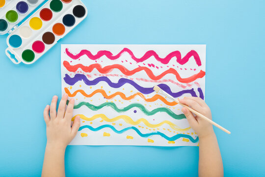 Baby boy hand holding paintbrush and drawing colorful wavy lines on white paper. Blue table background. Pastel color. Closeup. Child development. Learning painting. Point of view shot. Top down view.