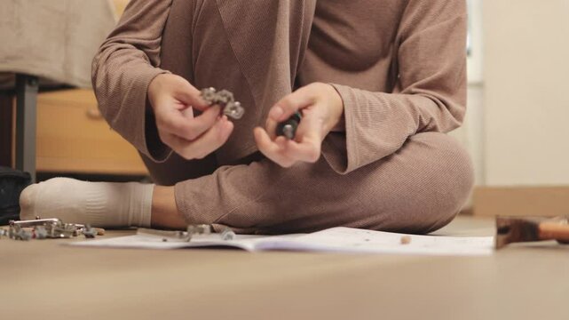 Home furniture assembly process showing focused woman sitting on floor, carefully constructing furniture piece using screwdriver, screws, and bolts while following detailed instructions