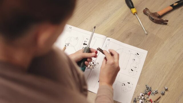 Woman carefully assembling furniture by screwing small metal parts together, following step-by-step instructions from an instruction booklet placed on a wooden floor