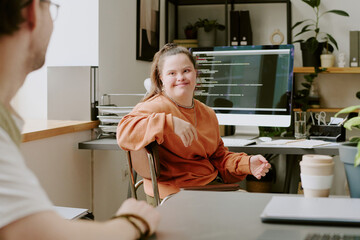 Wide shot of female programmer with down syndrome sitting on chair and looking at her male coworker