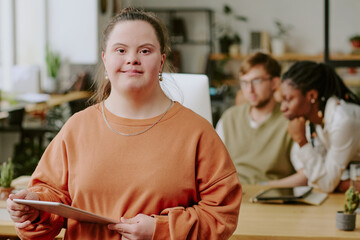 Medium close up portrait of smiling professional female programmer with down syndrome holding white tablet in hands while posing for photo in office © AnnaStills