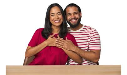 Seated young Latino couple at table has friendly expression, pressing palm to chest. Love concept.