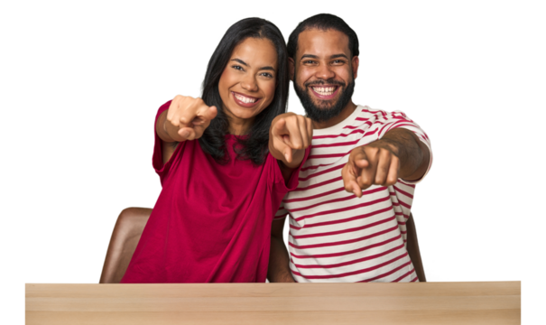 Seated young Latino couple at table cheerful smiles pointing to front.