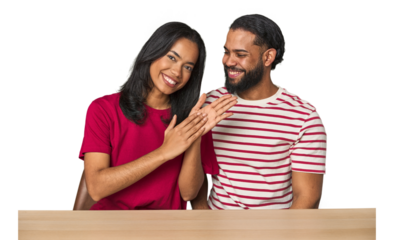 Seated young Latino couple at table feeling energetic and comfortable, rubbing hands confident.
