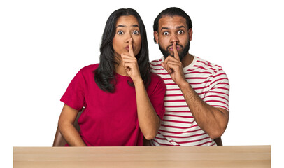 Seated young Latino couple at table keeping a secret or asking for silence.