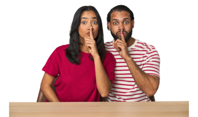 Seated young Latino couple at table keeping a secret or asking for silence.