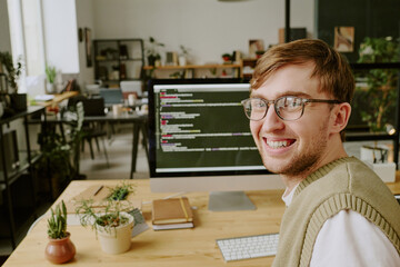 Medium close up of young adult man smiling while taking selfie with workplace background, desk with...