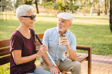 Elderly couple sitting on a bench in spring or summer park sunny day. Old blind woman with walking stick and man drinking coffee outdoors, life in retirement. Compassion caregiving concept