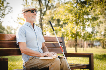 Side view of senior vision-impaired blind man sits on a park bench under sunlight, reading a book in braille. He is accompanied by a white cane and surrounded by lush green foliage in nature park