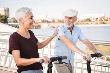 Active senior couple enjoying outdoor adventure with electric scooters and modern bridge. Eco life...