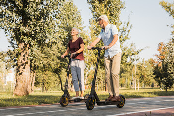Side view of senior man and woman elderly couple riding electric scooters on bridge under sunlight...