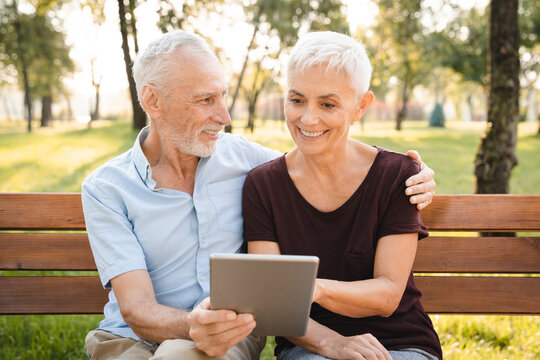 Caucasian senior elder couple sit on bench in park. Mature happy and enjoy with slow life. Old man and woman look at tablet and smile with life together. Retirement warm family lifestyle concept. - Powered by Adobe