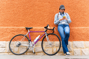 Young woman using smartphone leaning on orange wall with her racing bicycle