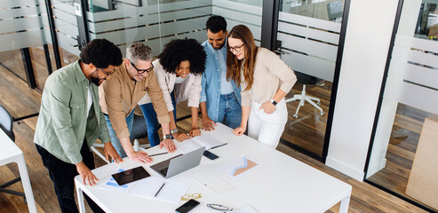 A diverse group of professionals collaborates in a modern office environment, brainstorming ideas over a laptop. They engage in teamwork, discussion, and strategy planning, showcasing productivity
