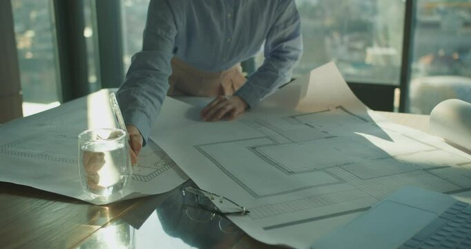 A detailed architectural plan being reviewed on a wooden table by a professional in a modern office, with sunlight reflecting off a glass of water and laptop nearby