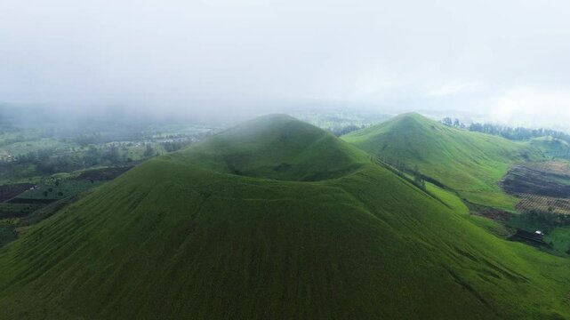 aerial footage of towering green hills surrounded by green meadows enveloped in a mystical fog. view of green hills peak with grassland and mountain views of Wurung Crater, Indonesia