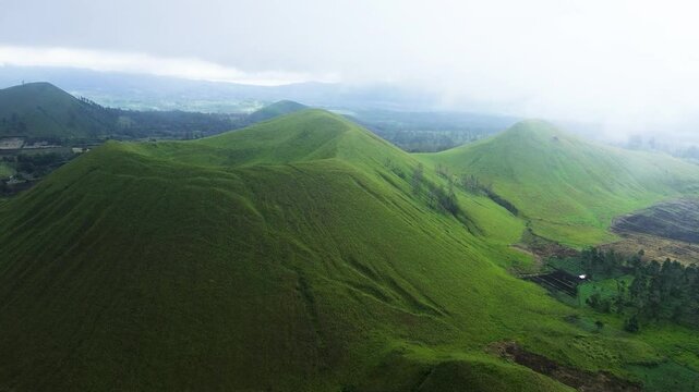 aerial footage of towering green hills surrounded by green meadows enveloped in a mystical fog. view of green hills peak with grassland and mountain views of Wurung Crater, Indonesia