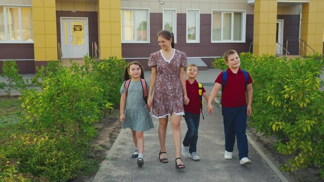 Mother leading children from school, female mother holding children hand, happy family, young nanny with children, schoolchildren with backpacks walking with aunt by the hand, little boys girl walking