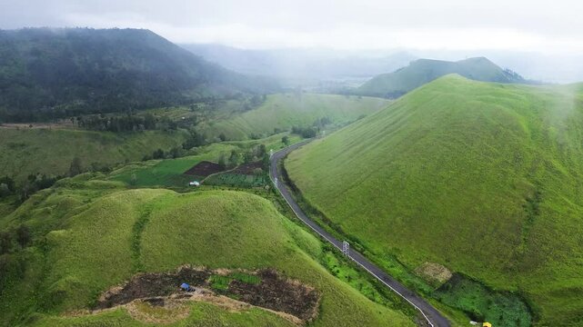 Stunning aerial footage tracking winding roads through vast green hills and geothermal power pipelines, surrounded by misty grasslands in Wurung Crater, Indonesia.
