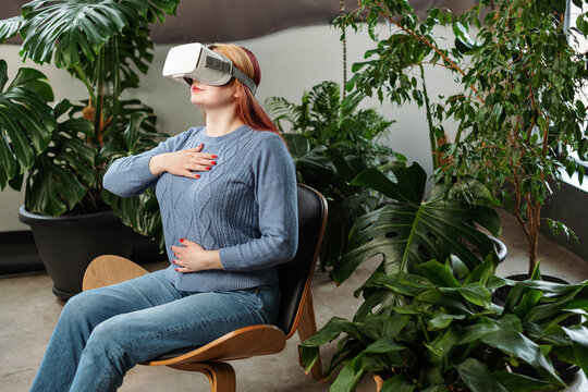 Young woman sitting on chair at green office workplace and using virtual reality headset while doing breath exercise during rest and meditation. Future technology concept.