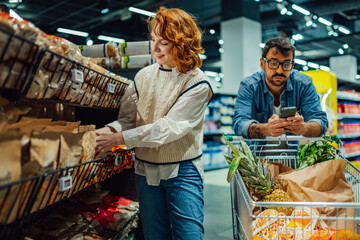Couple shopping for groceries at the supermarket, woman choosing products, man using smartphone
