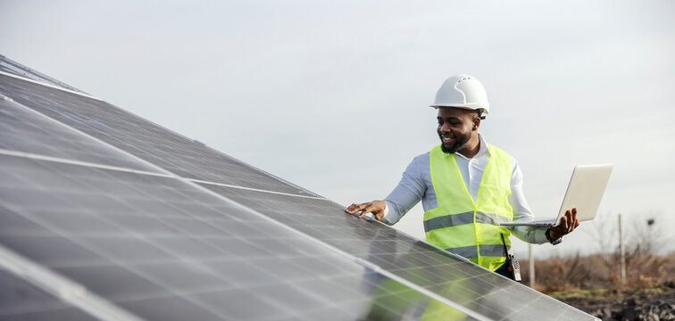 Multicultural electrical engineer standing at solar power station near solar panels and inspecting them while holding laptop.
