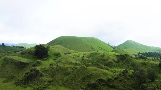 Stunning aerial footage of Kawah Wurung, Indonesia, showcasing towering green hills surrounded by expansive lush grasslands and green meadows.