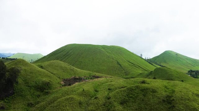 Stunning aerial footage of Kawah Wurung, Indonesia, showcasing towering green hills surrounded by expansive lush grasslands and green meadows.