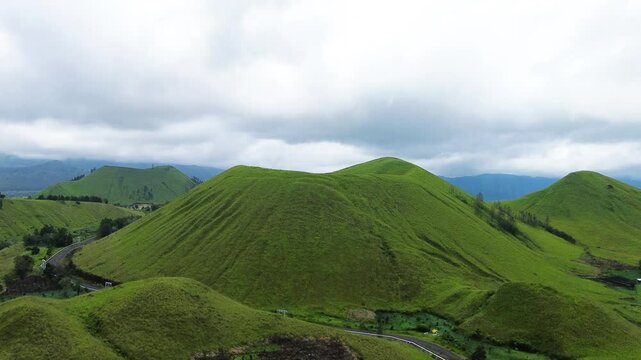 Stunning aerial footage of Kawah Wurung, Indonesia, showcasing towering green hills surrounded by expansive lush grasslands and green meadows.