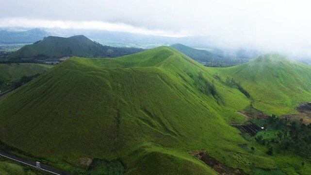 aerial footage of towering green hills surrounded by green meadows enveloped in a mystical fog. view of green hills peak with grassland and mountain views of Wurung Crater, Indonesia