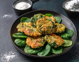 A plate of crispy spinach and cheese fritters is served on a bed of fresh spinach leaves, with a dark background and a sprinkle of flour.