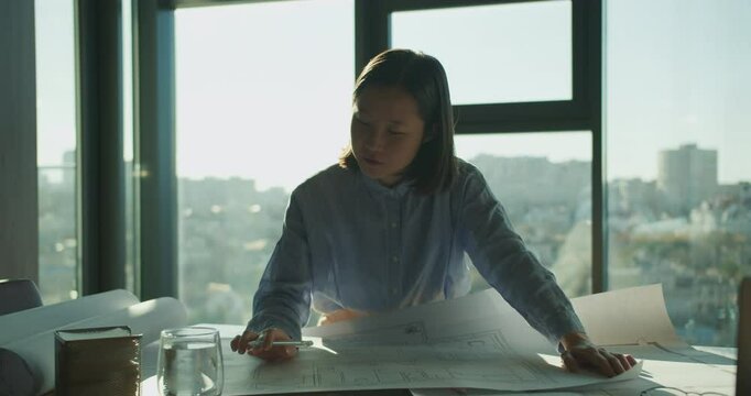 A young architect reviewing blueprints in a bright office with large windows and an urban skyline in the background, emphasizing creativity and precision