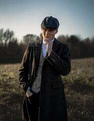 A stylish man in 1920s-inspired fashion, wearing a wool coat, vest, and flat cap, smokes a...
