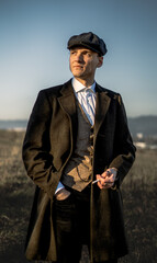 A stylish man in 1920s vintage fashion, wearing a wool coat, vest, and flat cap, stands in an open...