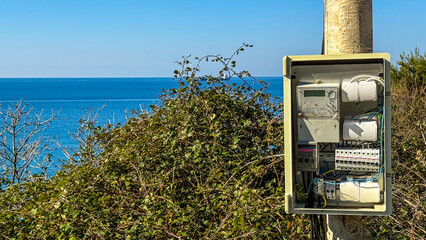 Electrical meter box beside coastal vegetation, utility installation along serene seashore,...