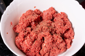 Seasoned raw ground meat in a scalloped white bowl with a coarse reddish texture. Ideal for making burgers, meatballs, or patties.