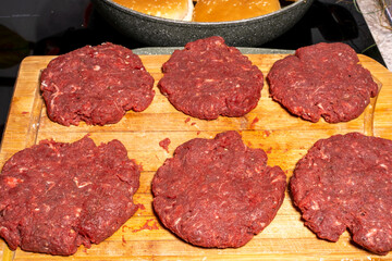 Uncooked burger patties on a wooden cutting board with hamburger buns being toasted in the...